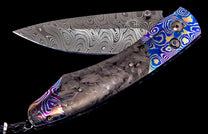 Folding knife with a Damascus blade, dark gray carbon fiber handle, and vibrant blue, gold, purple, and orange patterned metal accents.