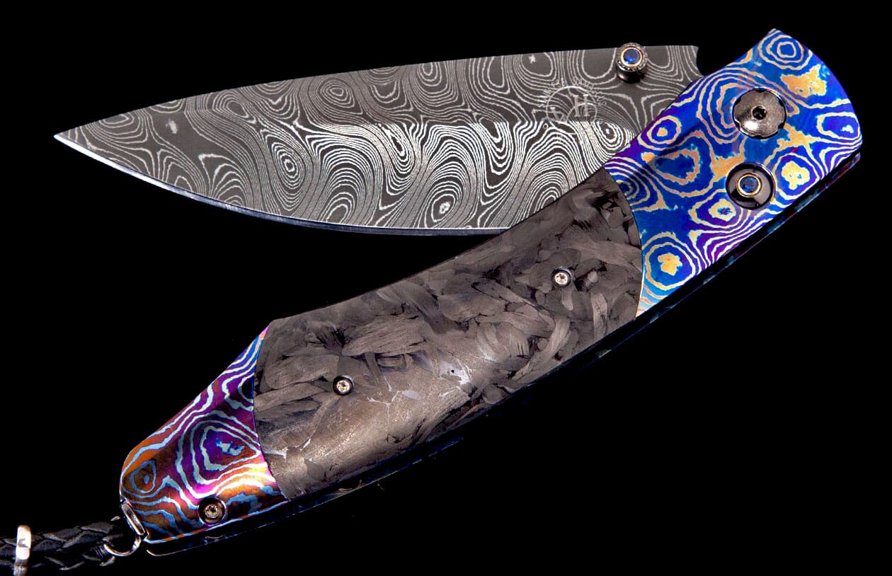 Folding knife with a Damascus blade, dark gray carbon fiber handle, and vibrant blue, gold, purple, and orange patterned metal accents.