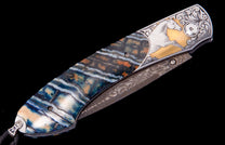 Ornate pocket knife with layered fossil handle in blue, white, and orange, featuring a gold inlay of a white horse, intricate scrollwork, and a patterned Damascus blade.