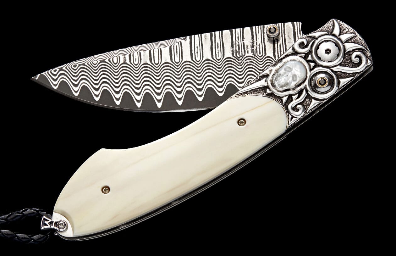 Folding knife with a wavy Damascus steel blade, cream handle, ornate silver bolster with skull carving, and black braided cord.