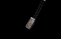 Silver-toned rectangular charm featuring an intricately carved skull design, attached to a braided black cord.