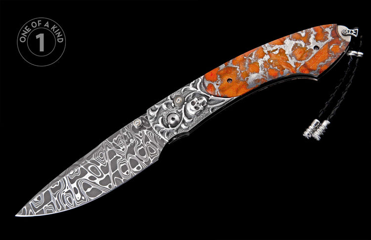 Highly decorative folding knife with a patterned Damascus steel blade, an ornate silver handle featuring a skull and orange-red inlay, and a black leather lanyard.