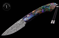 Ornate folding knife with a swirling Damascus steel blade and a multi-colored handle featuring abstract patterns, accented by a black braided lanyard.