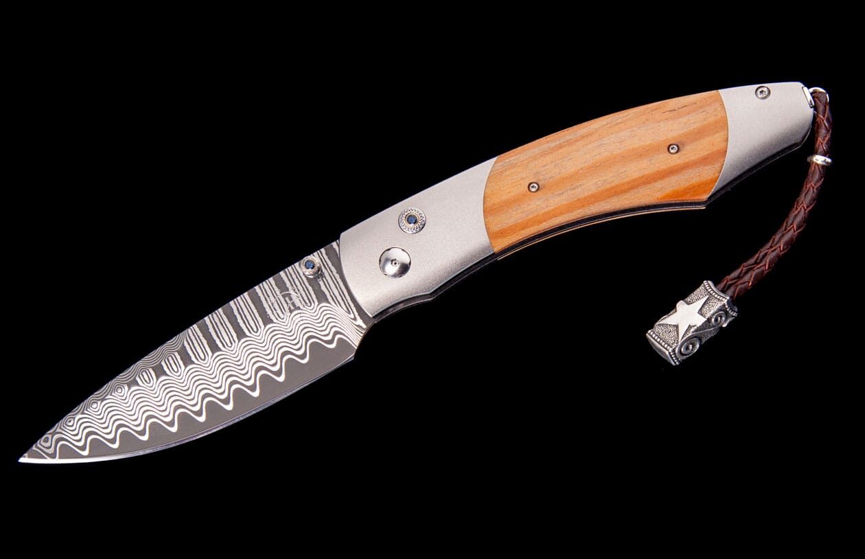 Folding knife with a patterned Damascus steel blade, light wood handle, and a braided leather lanyard with a decorative metal bead.