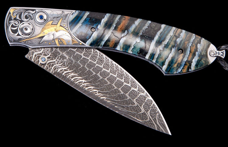Open decorative folding knife with a blue, brown, and white patterned handle, gold marlin engraving, blue gemstones, and a Damascus steel blade.