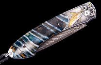 Folding knife with layered blue, black, and orange handle, ornate silver bolster featuring gold marlin, and patterned Damascus blade.