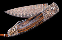 Custom folding knife with a wavy Damascus steel blade, a polished brown and blue handle, and ornate silver bolsters featuring bronze flame motifs and orange gemstones.