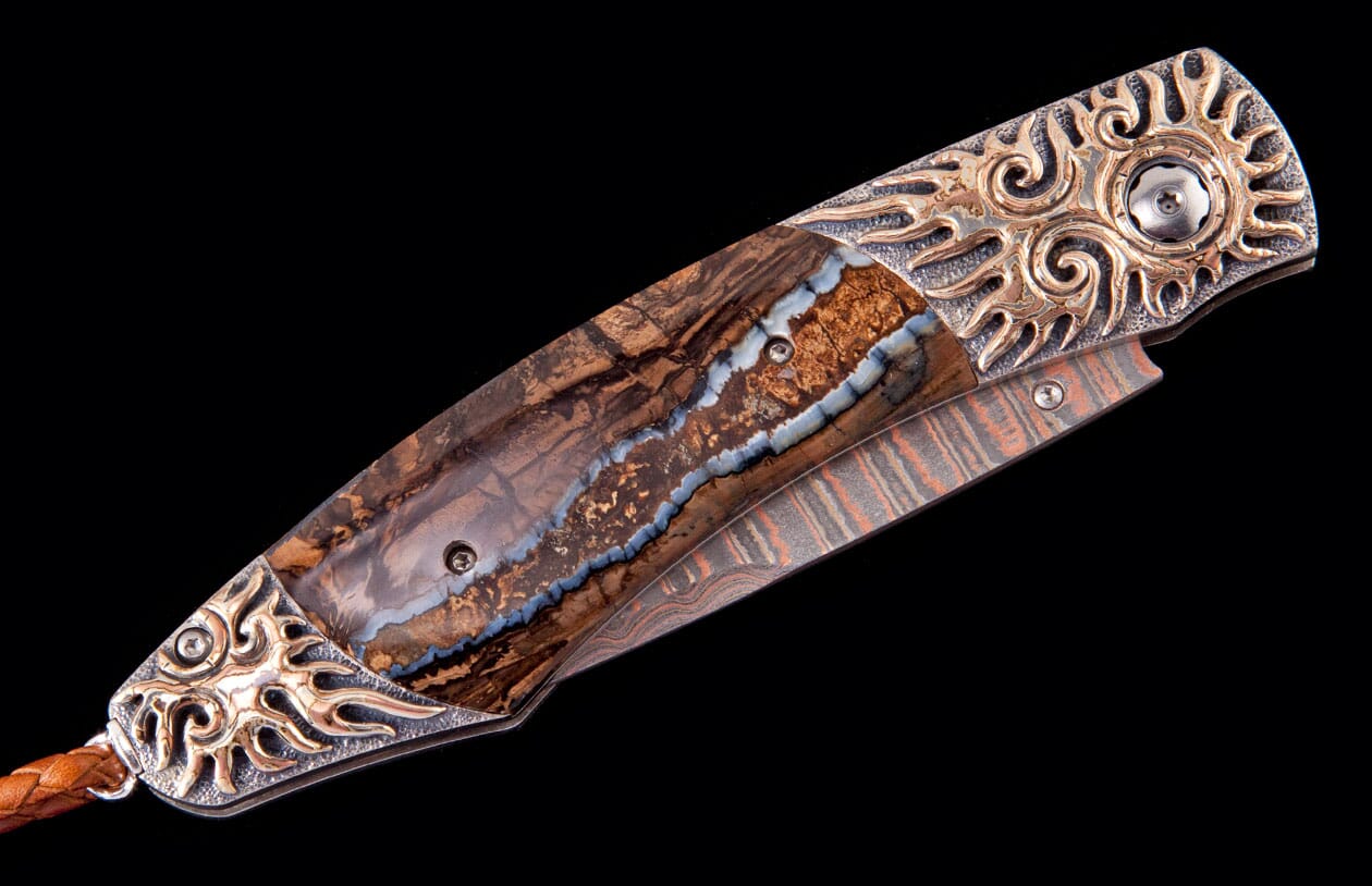 Ornate folding knife with a rich brown handle inlaid with blue opal, decorative bronze bolsters with sunburst motifs, and a Damascus steel liner.