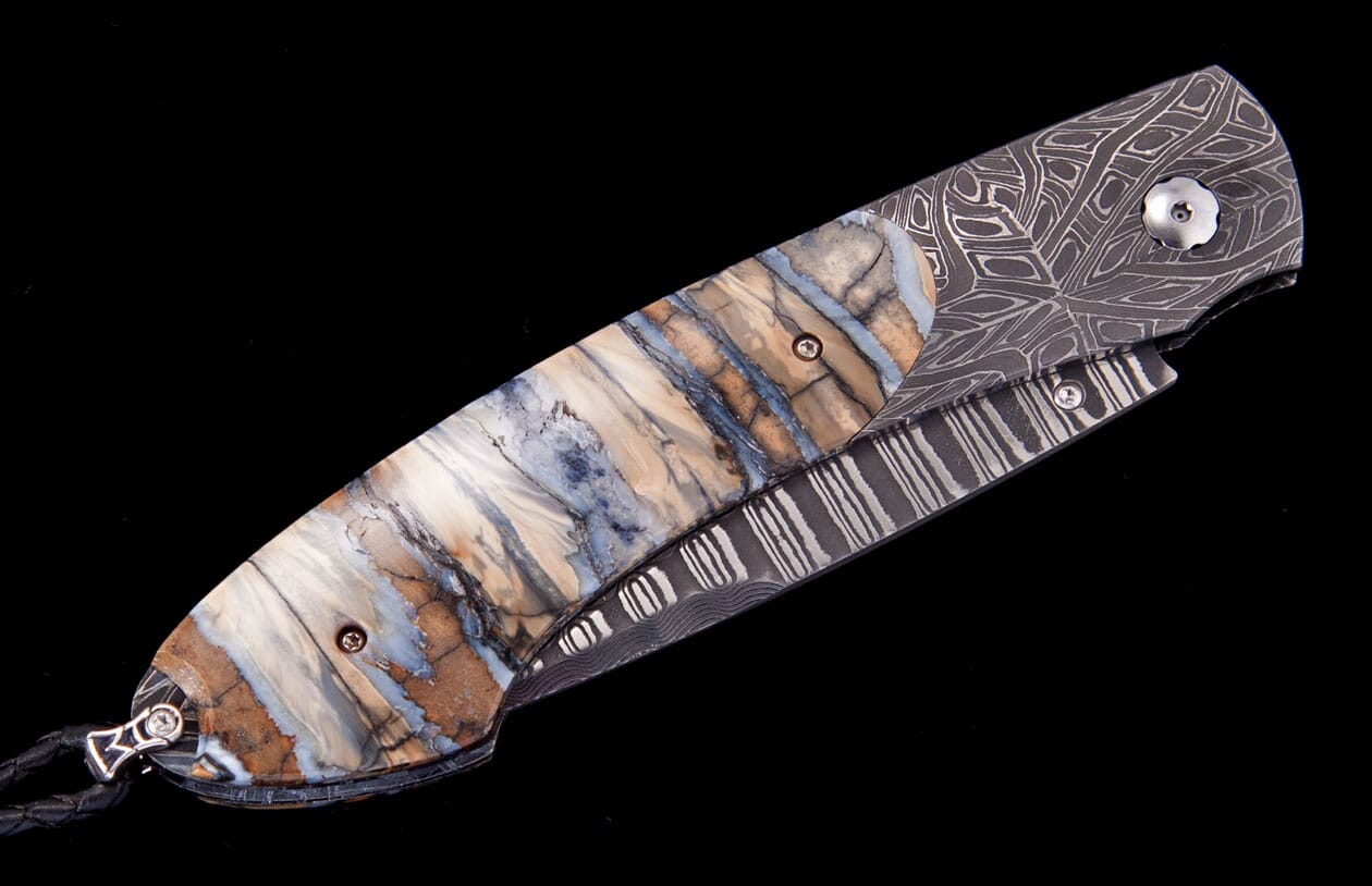 A folding knife with brown, white, and blue patterned handle scales by Tamreen, featuring intricate Damascus steel and a black leather lanyard.