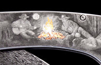 Western-themed engraved knife: two cowboys by an orange campfire under a moon, one playing guitar, with a patterned lower section.