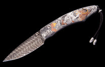 Western folding knife with Damascus blade, engraved handle showing silver, copper, and gold frontier scenes, and a leather lanyard.
