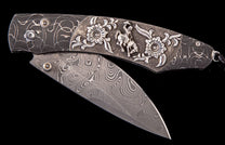 Damascus steel folding knife with a dark patterned handle featuring silver cowboy and floral embellishments.