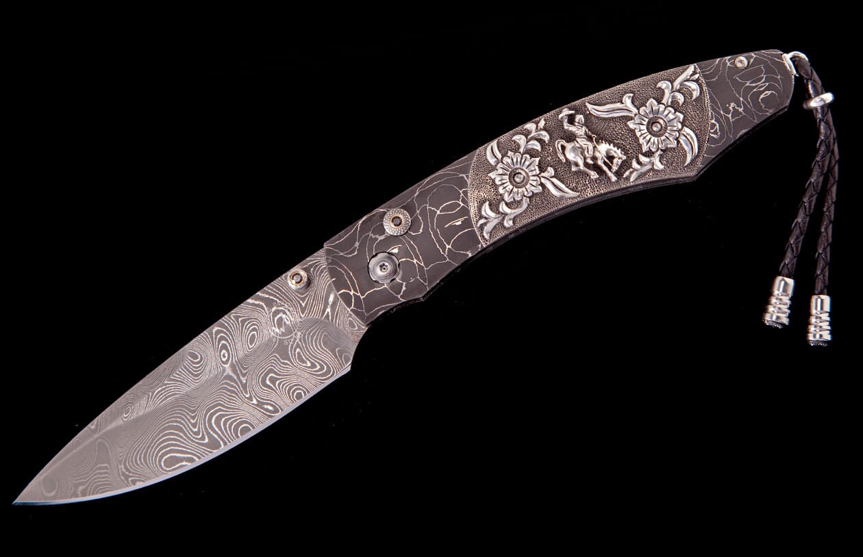 Damascus folding knife with an ornate handle featuring floral and cowboy engravings, and a braided leather lanyard.