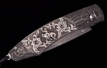 Dark Damascus steel folding knife with a silver-toned overlay featuring a cowboy on a bucking horse, floral patterns, and a crackle-patterned handle.