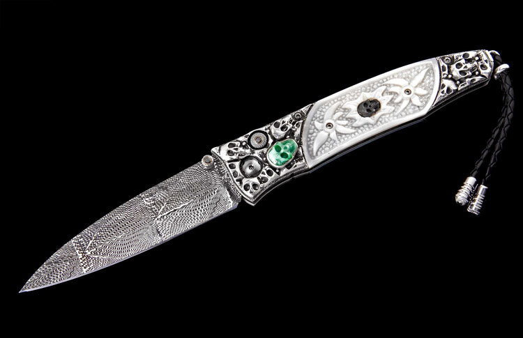 Ornate folding knife with a damascus blade, silver handle with carved skulls, white floral scales, and a green skull inlay.