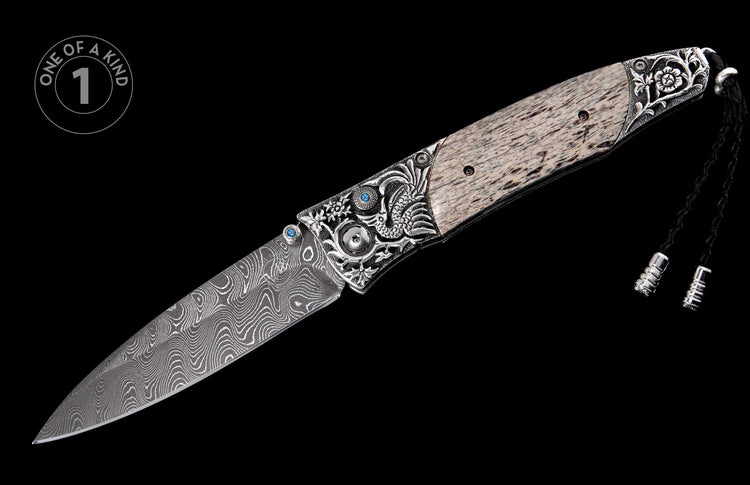 Folding knife with a wavy Damascus blade, ornate silver-tone bolsters with blue gemstones, a light patterned handle, and a black braided lanyard.