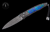 Ornate folding knife with a patterned Damascus blade, intricate handle featuring iridescent blue-green inlay, blue gemstone accents, and a braided black lanyard.