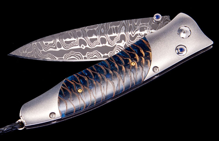 Folding knife with a patterned Damascus blade and a blue handle with a scaly gold design, featuring blue sapphire accents.