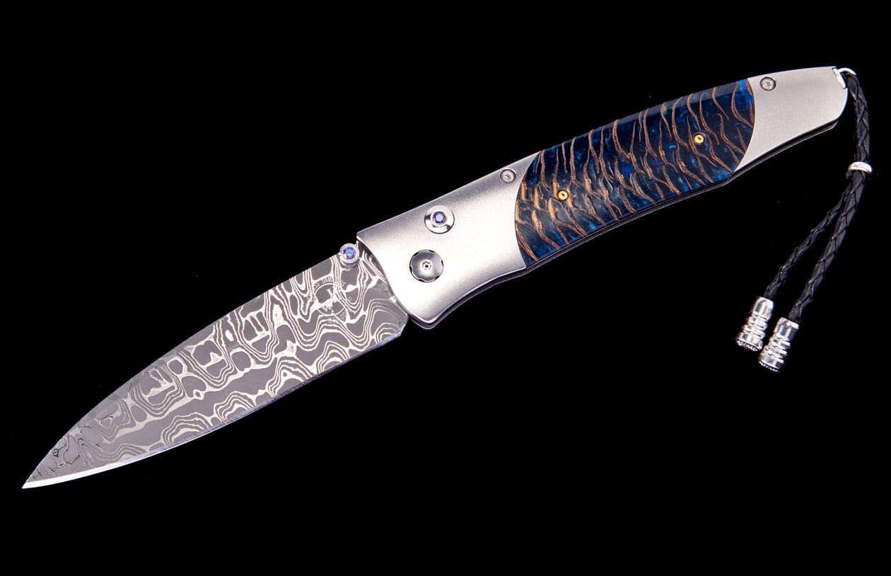 A custom folding knife by Tamreen, with a patterned Damascus blade, silver bolsters, a blue-streaked handle, and a black leather lanyard.