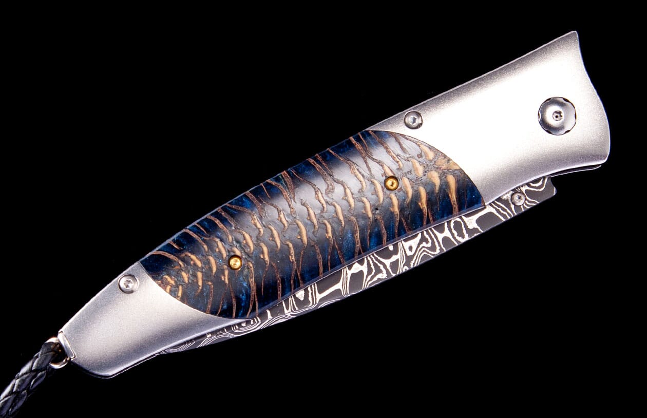 Silver-toned folding knife by Tamreen Custom with blue and brown scale-textured inlay, brass pins, Damascus blade, and braided leather lanyard.