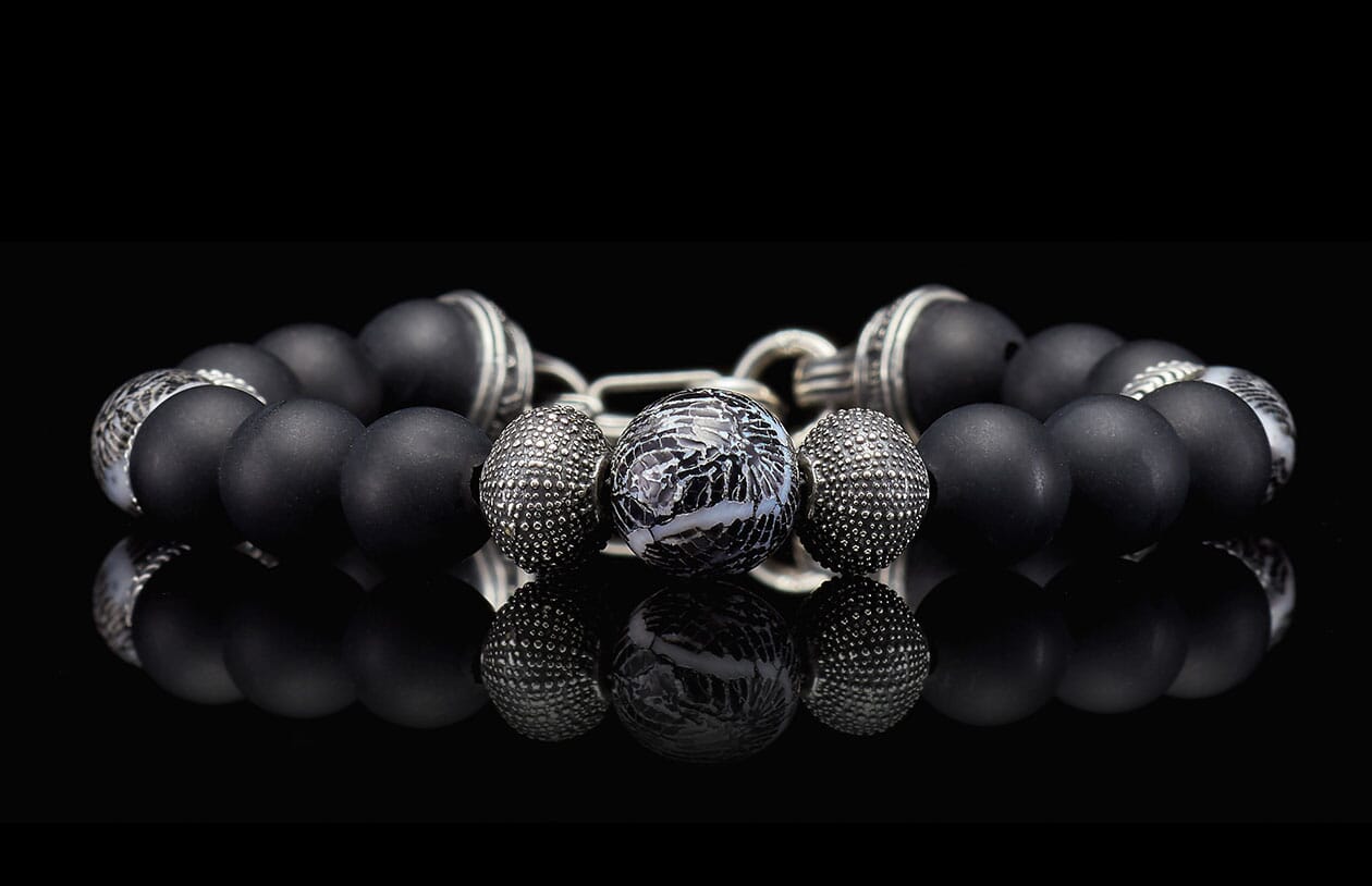Black matte beaded bracelet with a central marbled bead and textured silver spacers, reflecting on a dark surface.