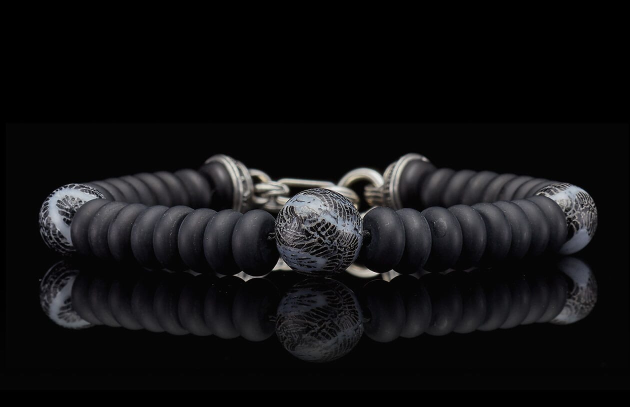 Black beaded bracelet with matte black and patterned grey beads, silver accents, reflecting on a dark surface.