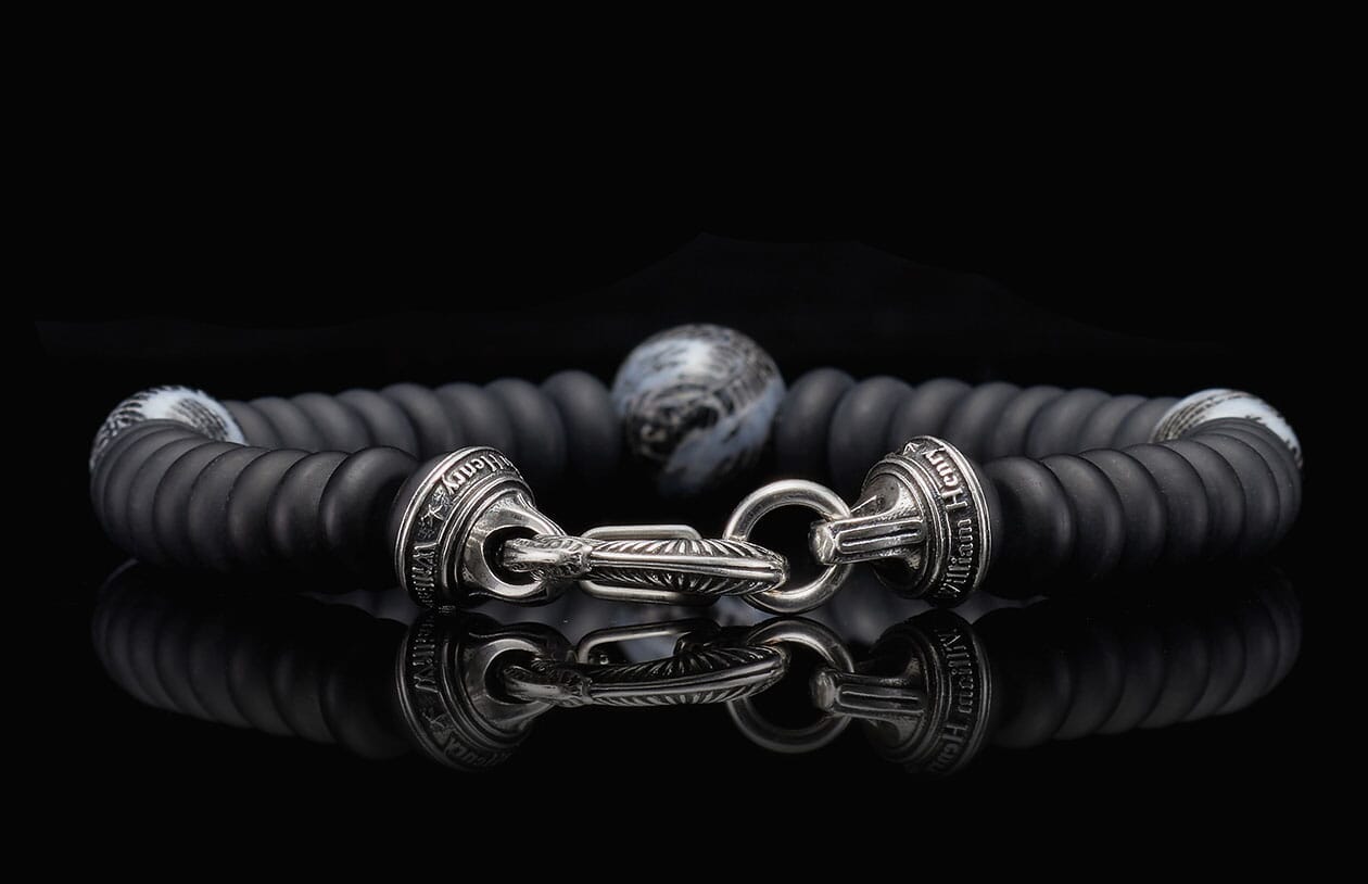 Matte black beaded bracelet with an ornate silver-tone clasp featuring engravings, a central patterned bead, and a strong reflection.