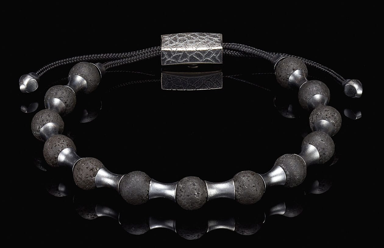 Lava stone bracelet with dark porous beads, silver-toned metal spacers, and a hammered rectangular clasp on a black reflective surface.