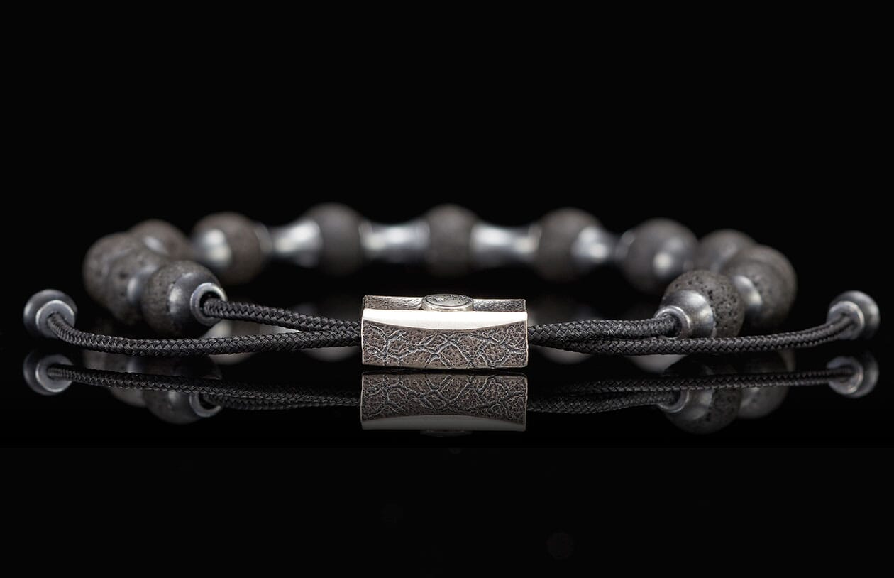 Dark gray lava stone beaded bracelet with a textured silver-toned metal clasp and adjustable black braided cords.