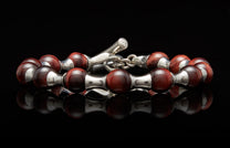 Deep reddish-brown beaded bracelet with polished silver spacers and toggle clasp, reflected on a dark surface.