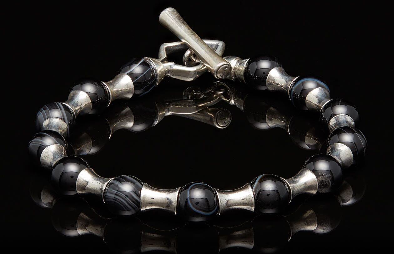 Silver and black beaded bracelet with a toggle clasp, featuring striped agate and smooth black beads separated by silver spacers on a reflective surface.