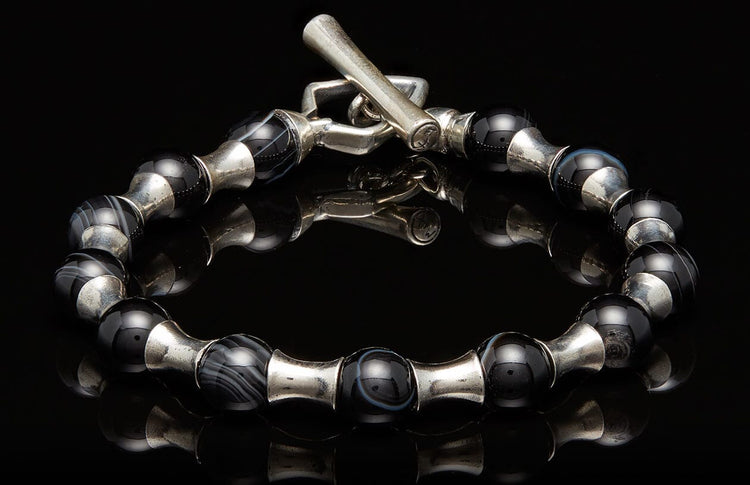 Silver and black beaded bracelet with a toggle clasp, featuring striped agate and smooth black beads separated by silver spacers on a reflective surface.