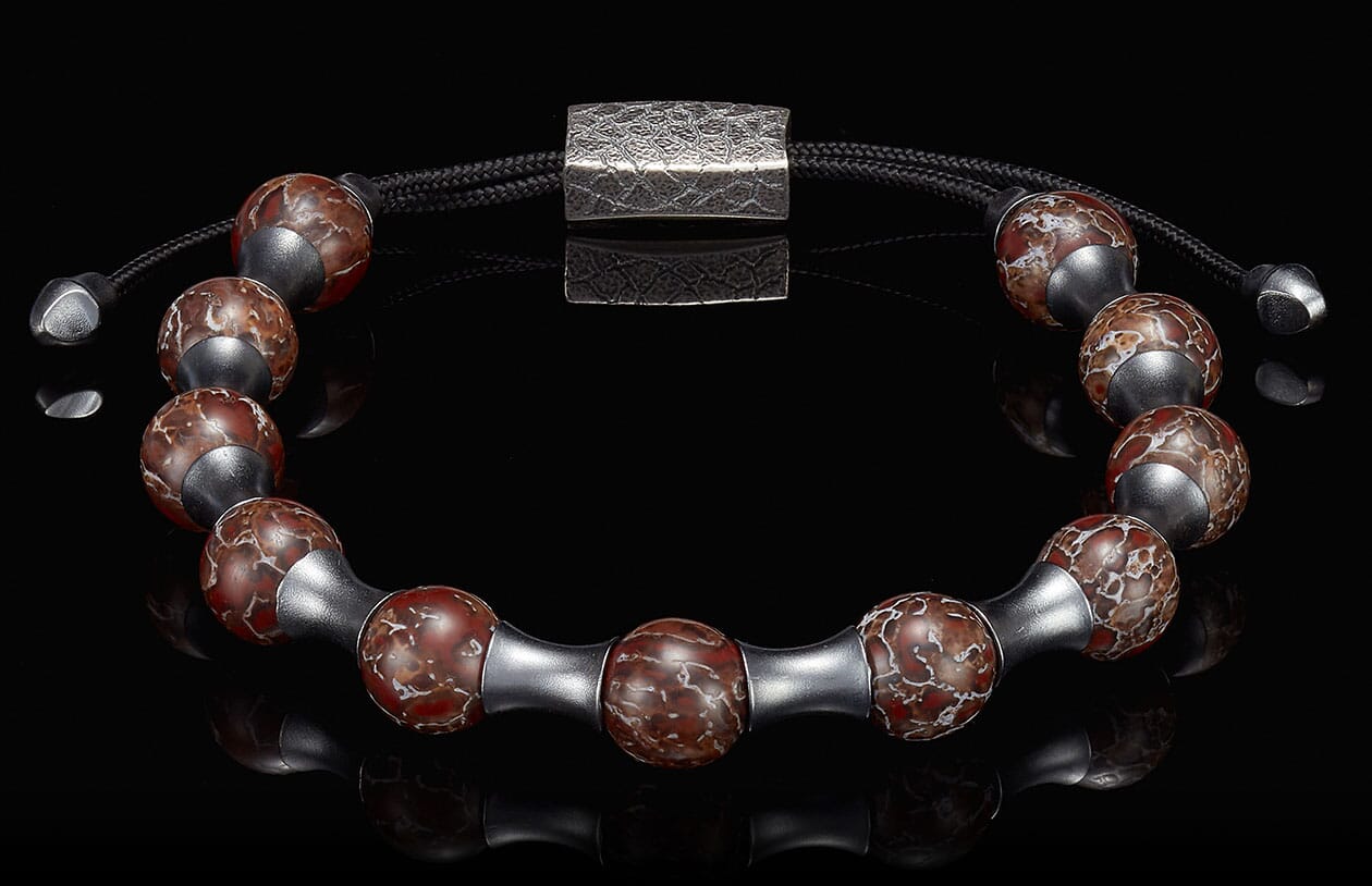 Bracelet with reddish-brown marbled stone beads, silver-toned cylindrical spacers, black braided cord, and textured rectangular clasp.