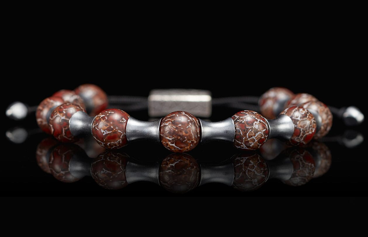 Beaded bracelet with red-brown marbled beads, silver tube spacers, and a central rectangular clasp, reflected on a dark surface.