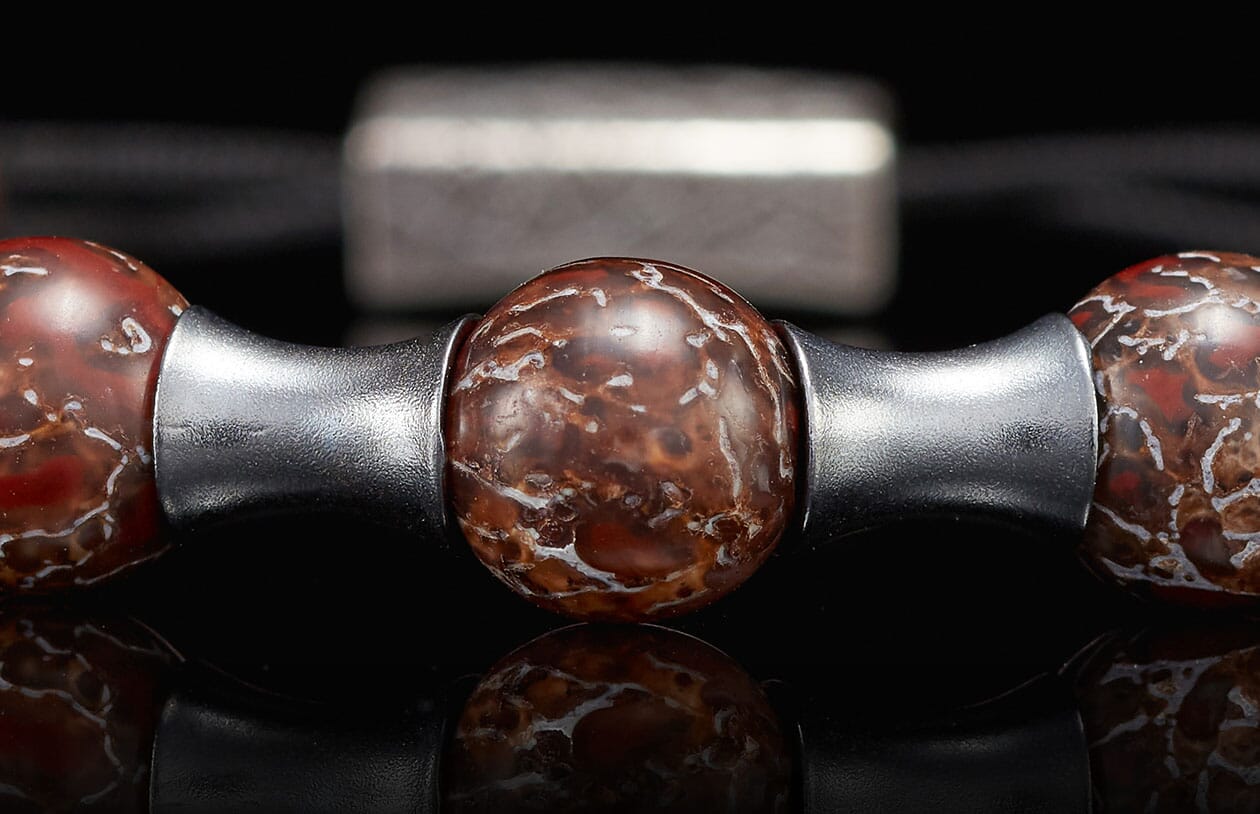 Reddish-brown stone beaded jewelry with white fossil-like patterns and silver metallic spacers, reflected on a dark surface.