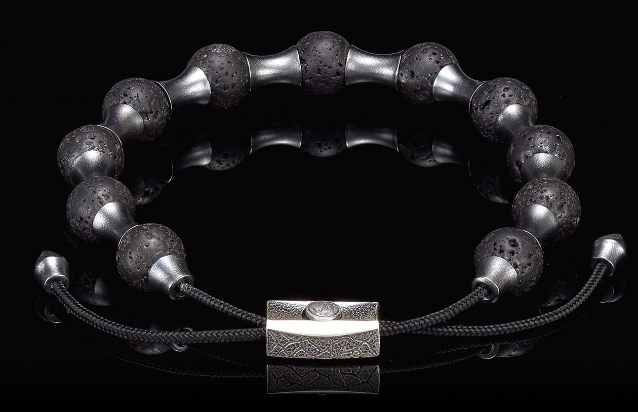 Black lava stone and silver-toned metal bracelet with textured beads and a rectangular patterned clasp on a reflective black surface.