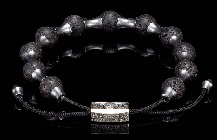 Black lava stone and silver-toned metal bracelet with textured beads and a rectangular patterned clasp on a reflective black surface.