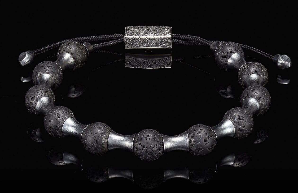 Black lava stone and silver-tone metal beaded bracelet with a textured rectangular clasp, reflected on black.