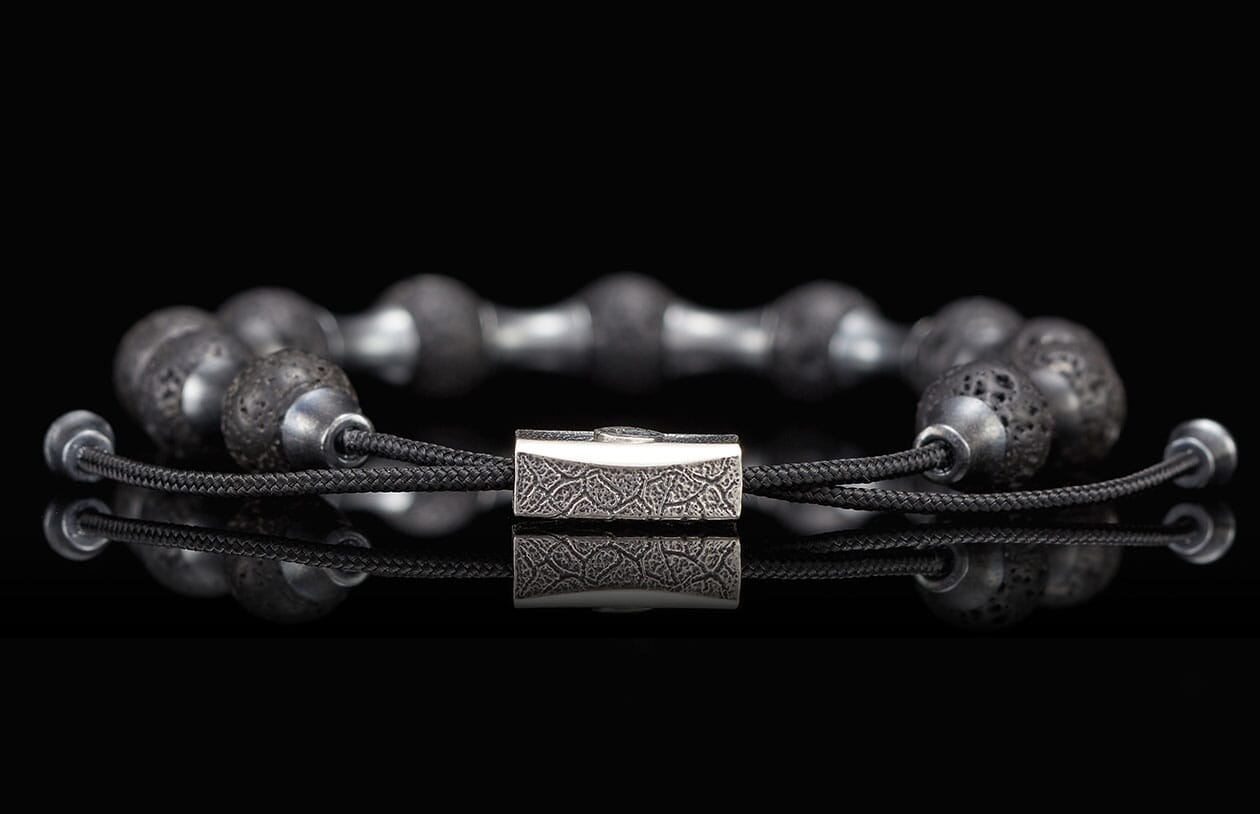 Black lava stone bracelet with an intricately engraved silver clasp, reflected on a glossy surface.