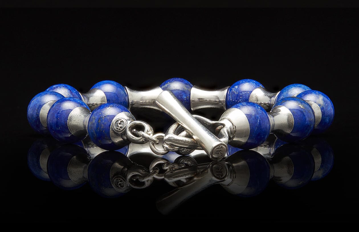 Blue lapis lazuli and silver beaded bracelet with a T-bar clasp, reflecting on a dark surface.