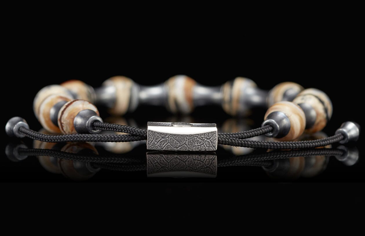 Braided black cord bracelet with alternating light brown patterned beads and metallic beads, featuring a textured rectangular silver clasp, reflected on a black surface.