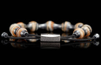 Braided black cord bracelet with alternating light brown patterned beads and metallic beads, featuring a textured rectangular silver clasp, reflected on a black surface.