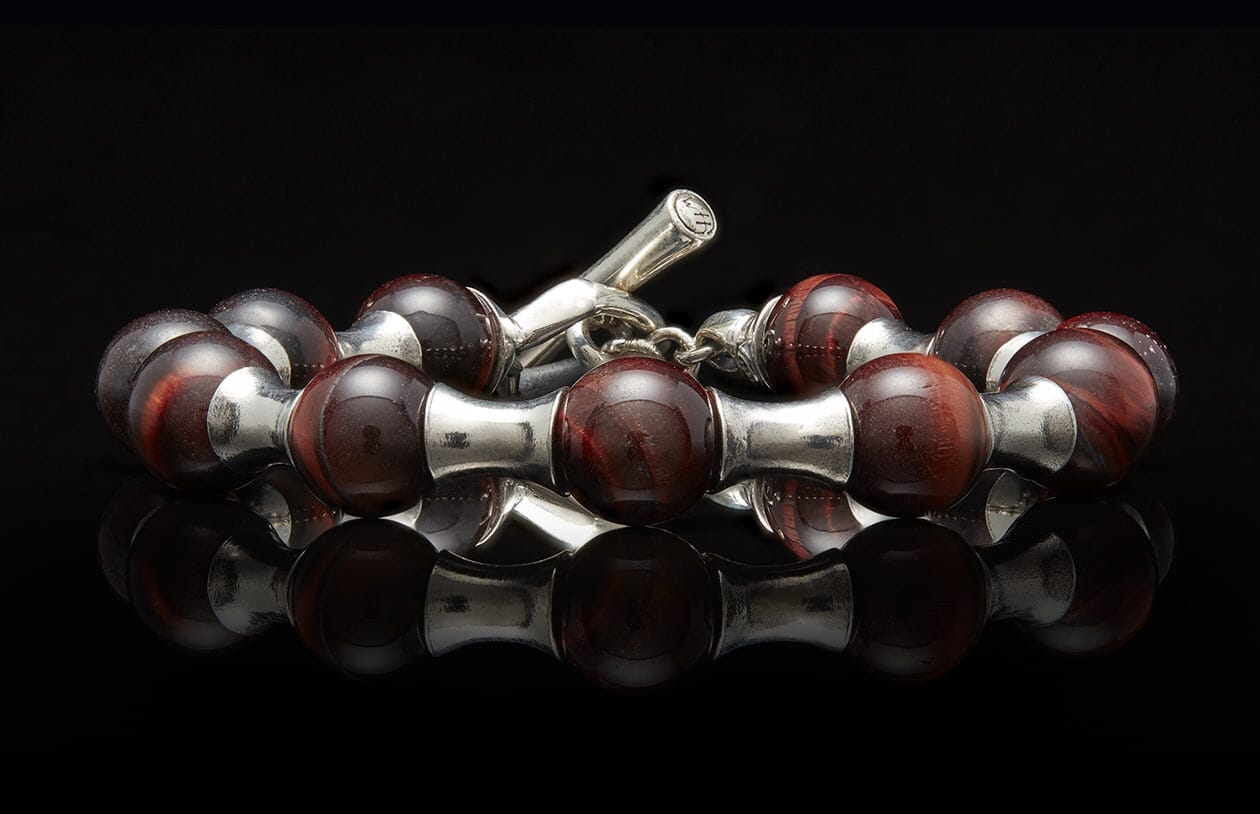 Polished reddish-brown beaded bracelet with chatoyant streaks and silver accents, featuring a toggle clasp, reflected on a black surface.