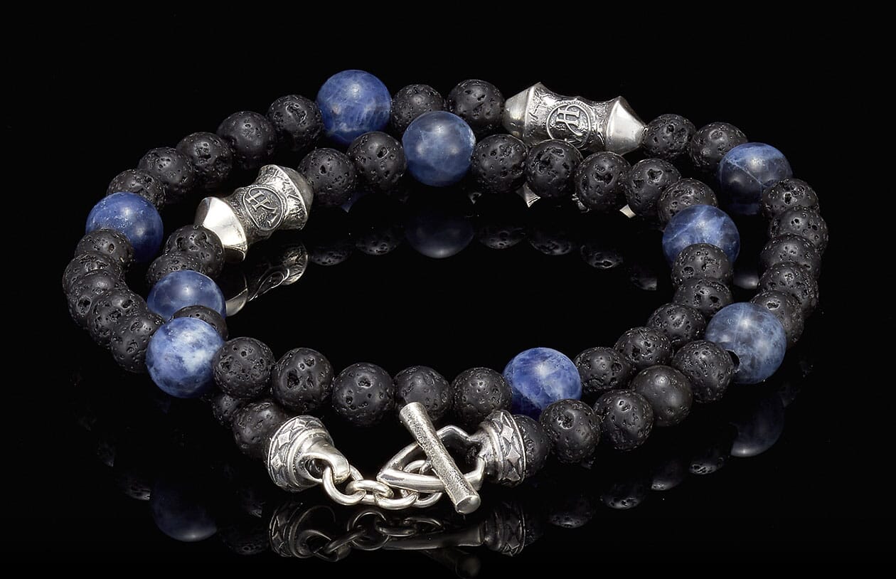 Sodalite tether made of porous black lava stone and marbled blue sodalite beads, featuring intricate silver-toned accent beads and a toggle clasp, displayed on a reflective black surface.