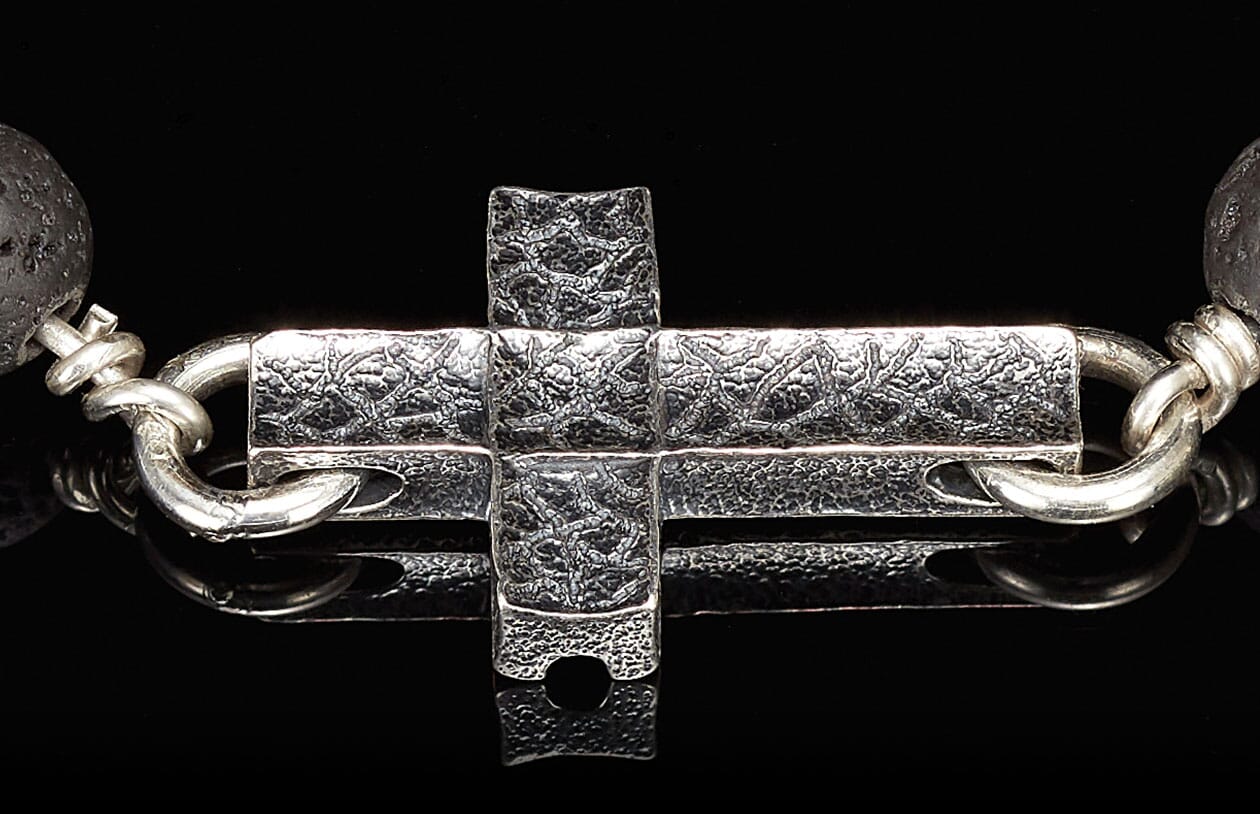 Cross-shaped silver jewelry clasp with textured surface, connected to a dark porous lava bead.