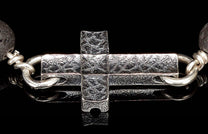 Cross-shaped silver jewelry clasp with textured surface, connected to a dark porous lava bead.