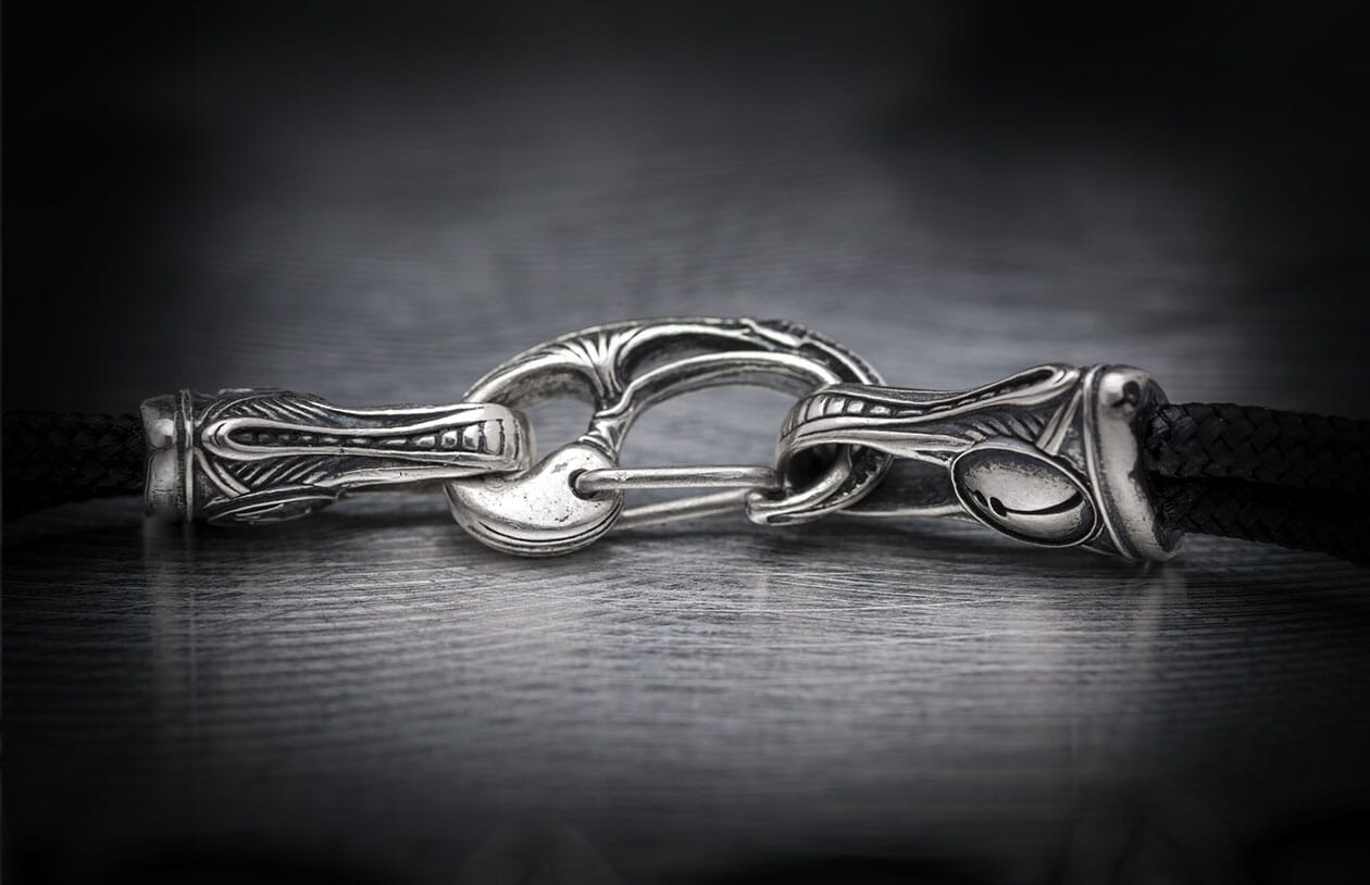 Silver jewelry clasp featuring elaborate decorative metalwork, connected to a black braided cord.