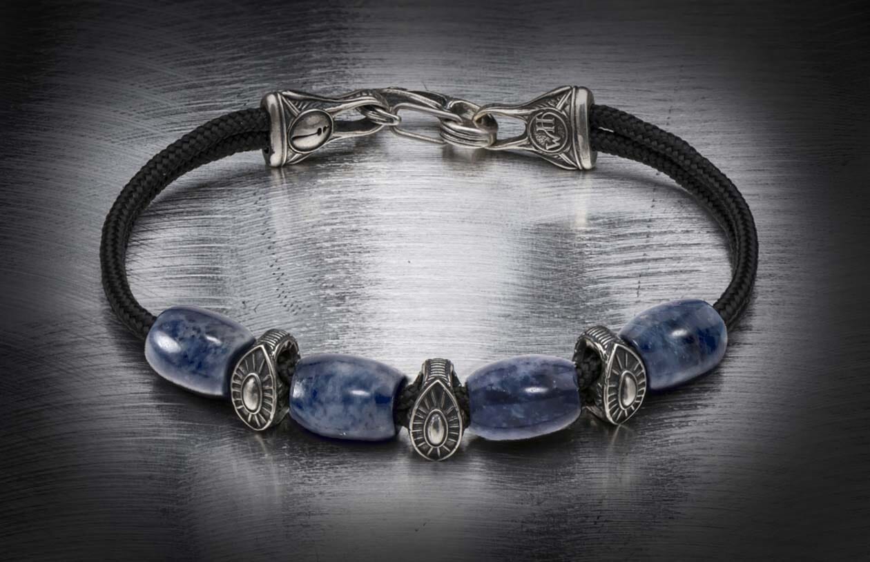 Blue sodalite and black cord bracelet with ornate silver-toned metal beads and clasp on a dark brushed surface.