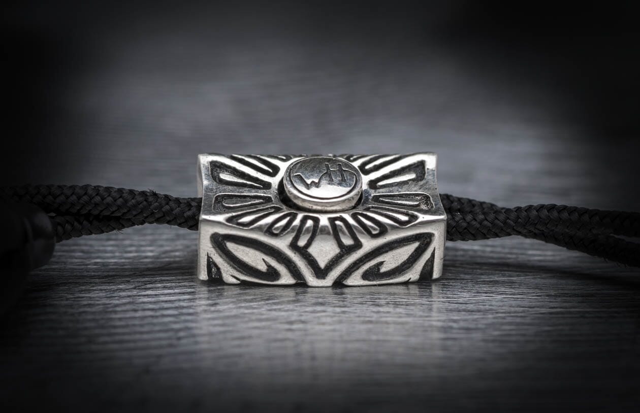 Silver rectangular metal bead with ornate carvings and a raised oval 'WH' logo, threaded on a black braided cord.