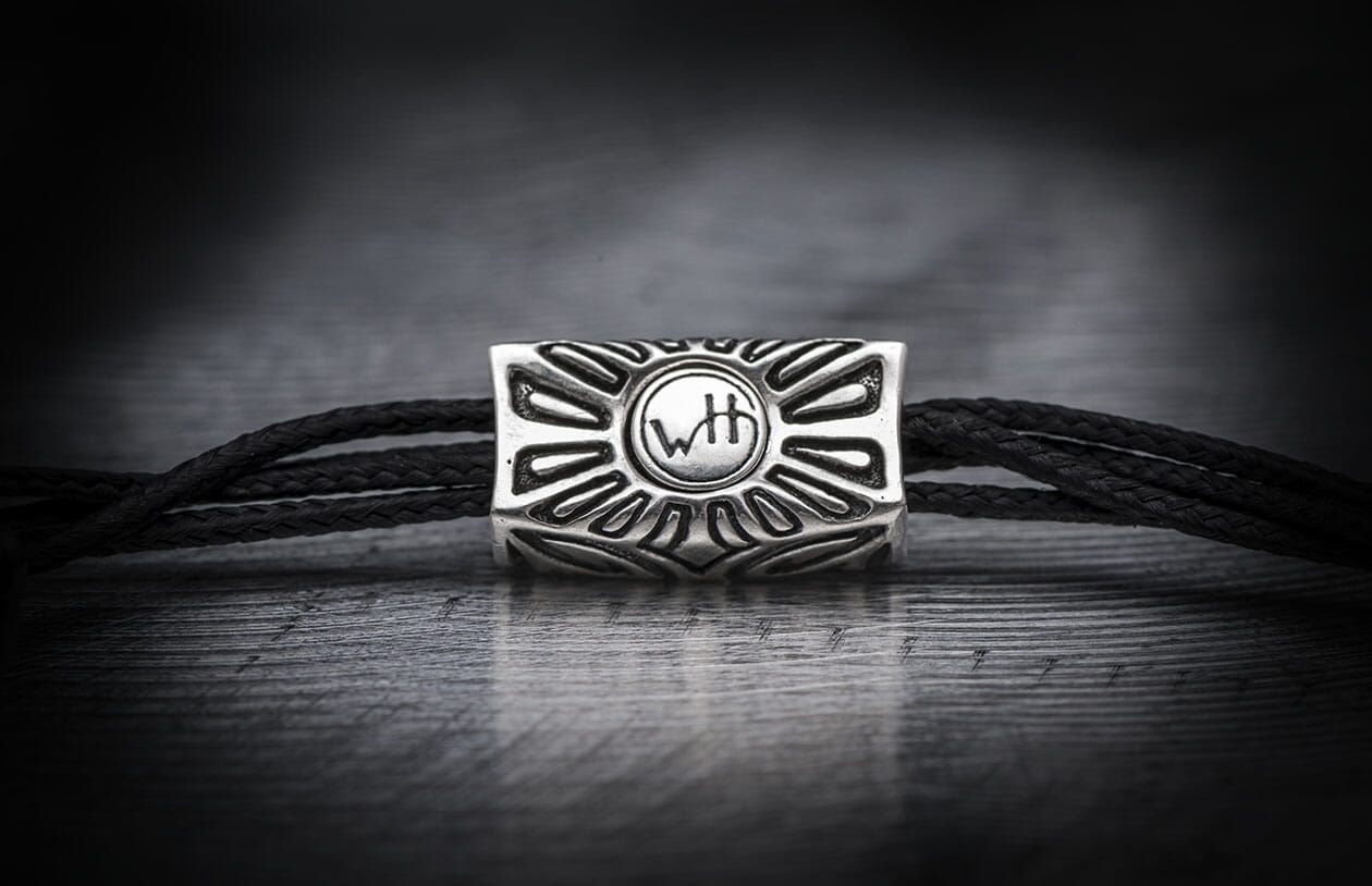 Black cord bracelet with a silver-toned metal bead featuring a carved sunburst design and central 'WtH' initials.